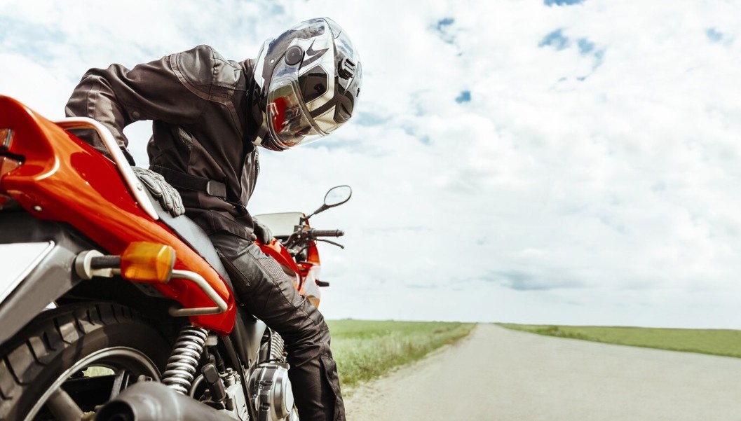 Motorcyclist in full gear sitting on a red bike, looking down, stranded on an empty road under a cloudy sky.
