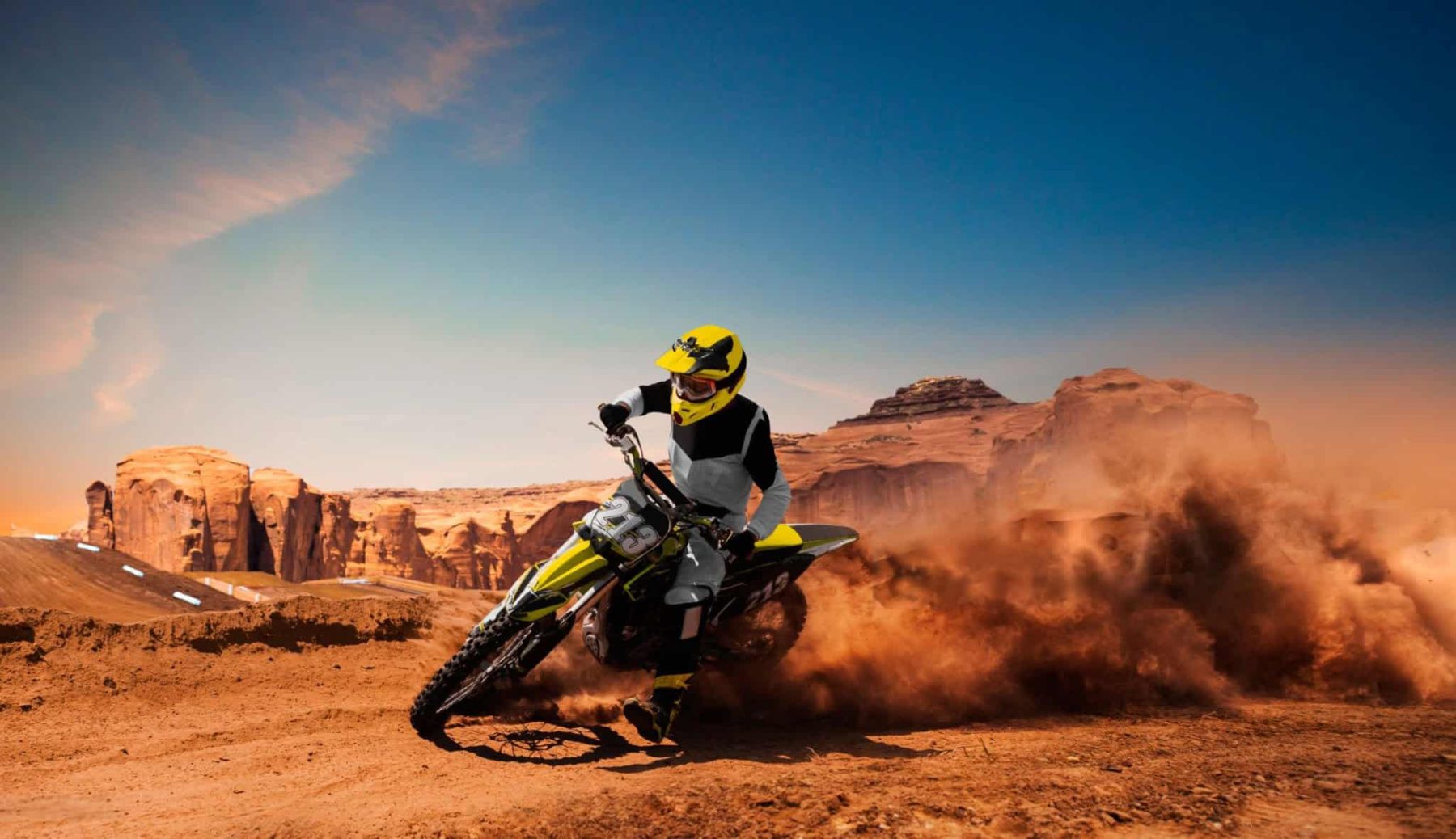 Dirt bike rider carving a fast turn in a desert trail, kicking up a large cloud of dust with dramatic motion and rocky terrain in the background.