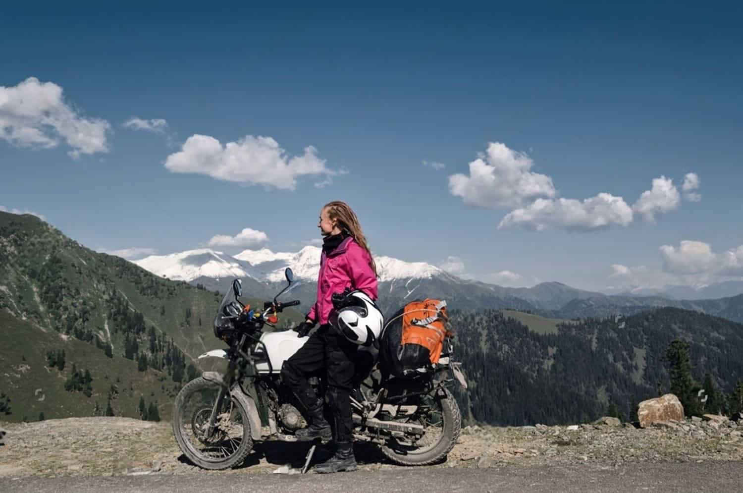 Woman in pink motorcycle jacket standing beside adventure bike in mountain setting, challenging the idea that color equals quality for women’s gear.