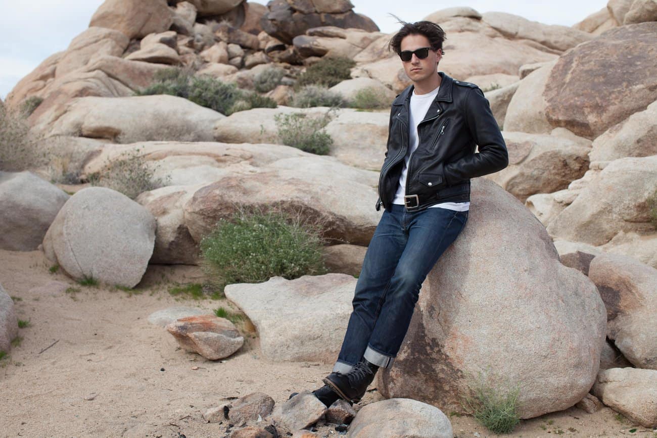 Man wearing a classic Schott NYC leather motorcycle jacket and jeans, leaning on a boulder in a desert landscape