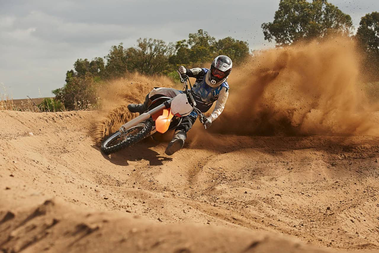 Motocross rider aggressively cornering on a dirt track, kicking up a massive dust cloud in a high-speed turn.