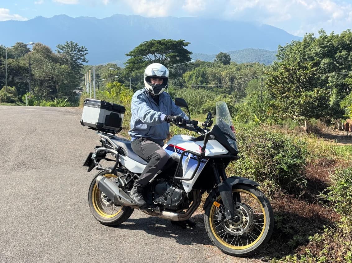 Adventure rider wearing summer motorcycle pants and gear on a Honda Transalp, parked on a rural road with forested mountains in the background