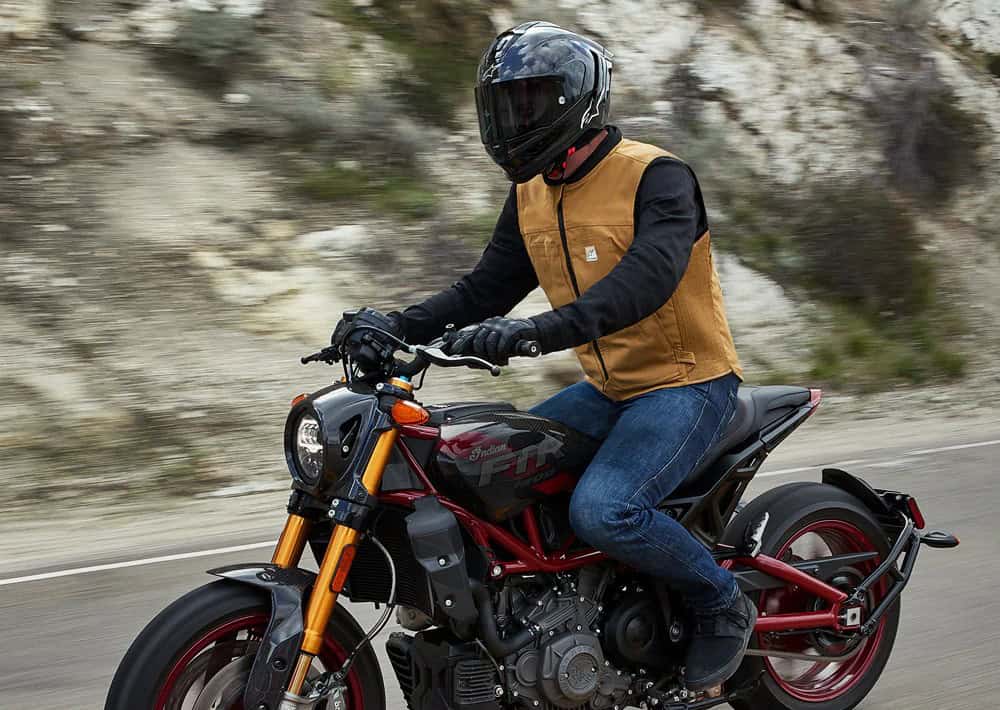 Motorcyclist wearing a tan airbag vest and black helmet riding on a mountain road, illustrating real-world protective gear in use.
