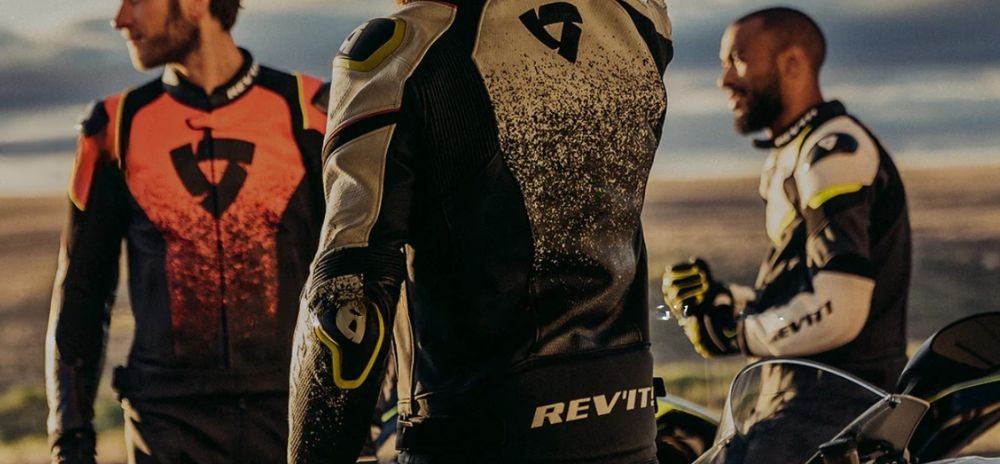 Three motorcyclists wearing REV'IT! riding gear, standing near bikes at sunset