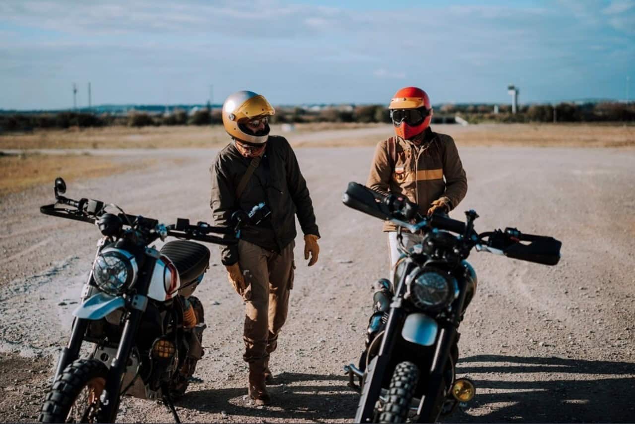 Two motorcyclists wearing helmets and textile riding gear standing beside scrambler-style bikes in a sunny, open gravel area