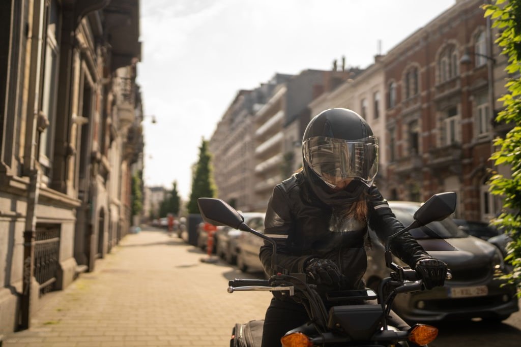 Motorcyclist in black gear rides through an urban street, wearing gloves and a full-face helmet.
