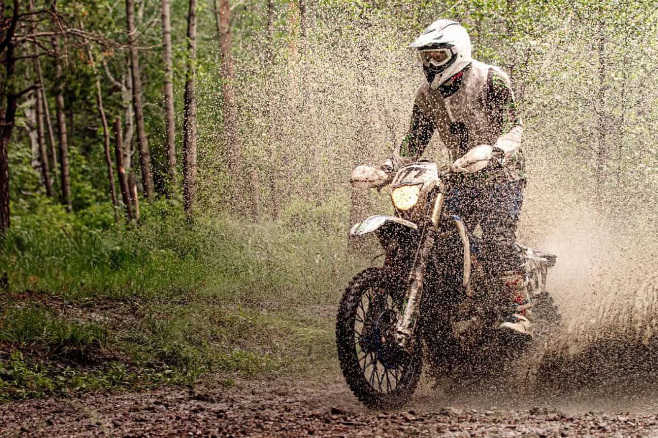 Dirt bike rider splashing through a large muddy puddle in a forest trail, water spraying up in all directions in wet riding conditions.