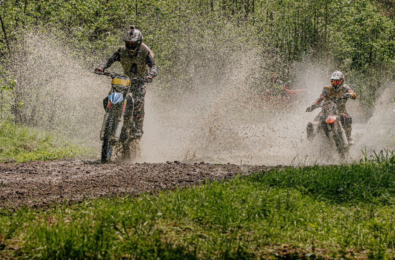 Two riders splashing through muddy water on off-road motorcycles, highlighting the importance of waterproof gear in wet conditions.