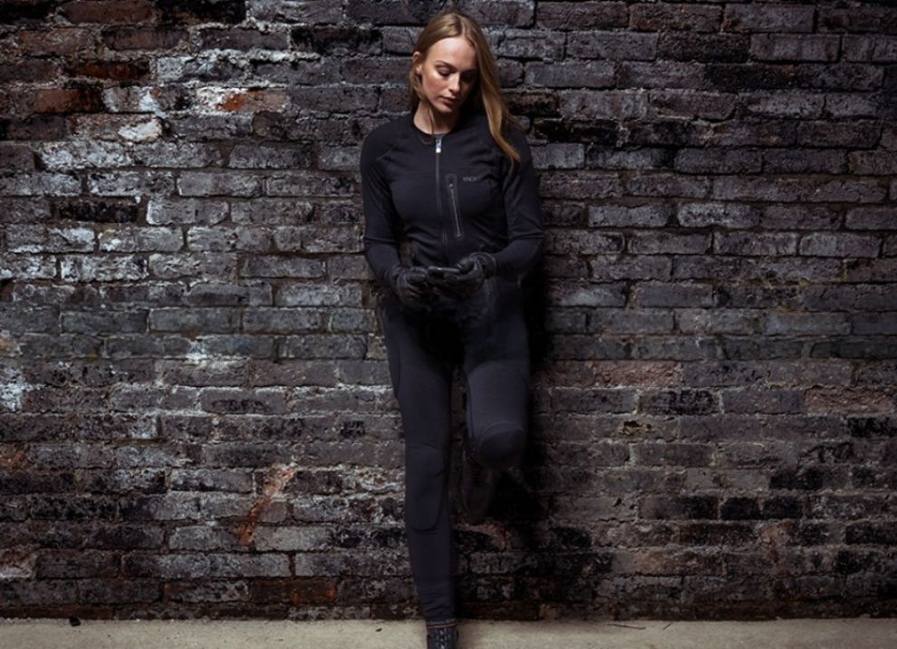 Woman wearing fitted motorcycle gear and leather gloves, standing against a brick wall