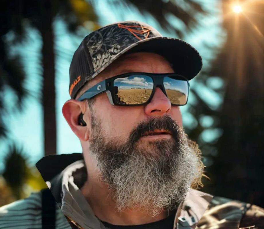 Man wearing a cap, beard, sunglasses, and Decibullz earplugs outdoors