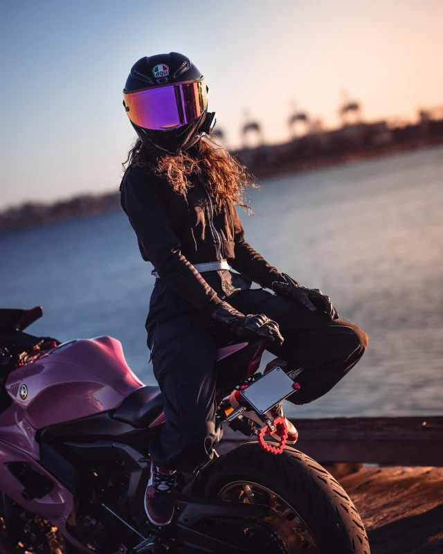 Motorcyclist in a black outfit and mirrored AGV helmet sitting on a pink sportbike by the water at sunset.