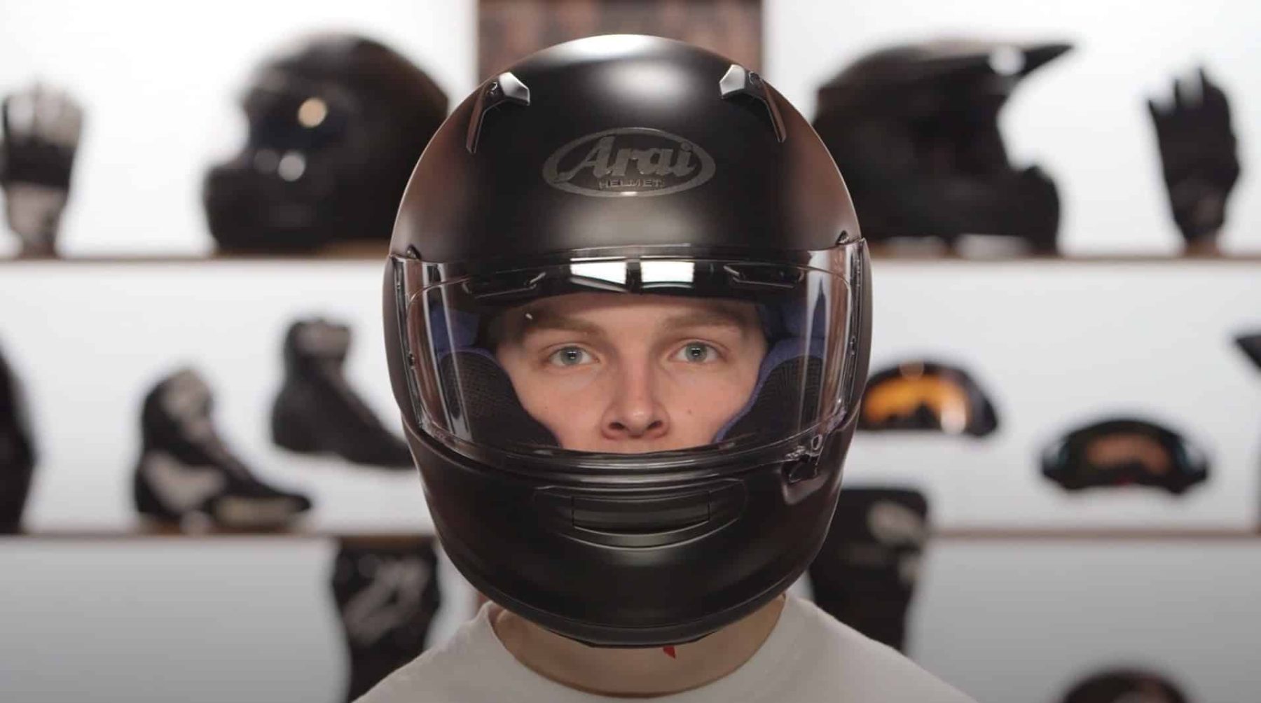 Front view of a rider wearing a matte black Arai Regent-X helmet with clear visor, photographed indoors against a gear wall.