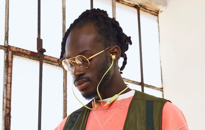 Man wearing neon yellow Beats Flex wireless earphones with a neckband, standing indoors by a window in casual attire.