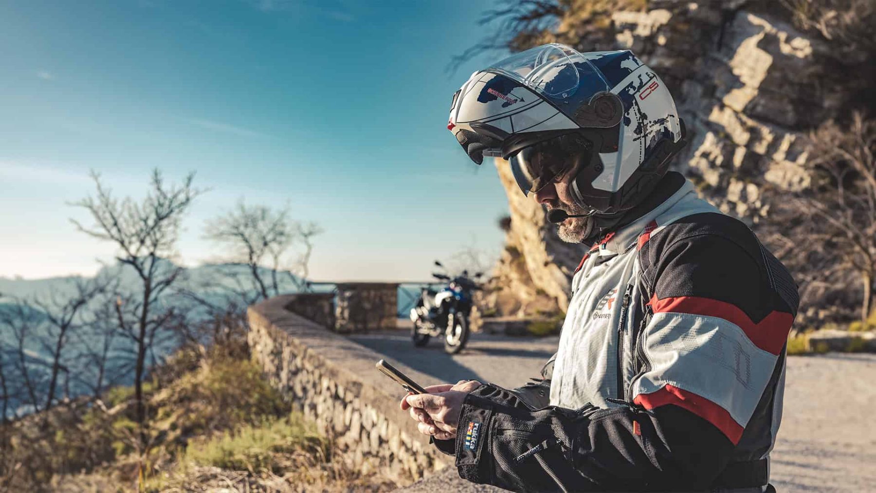 Touring rider in modular helmet using phone, bike parked by scenic mountain road