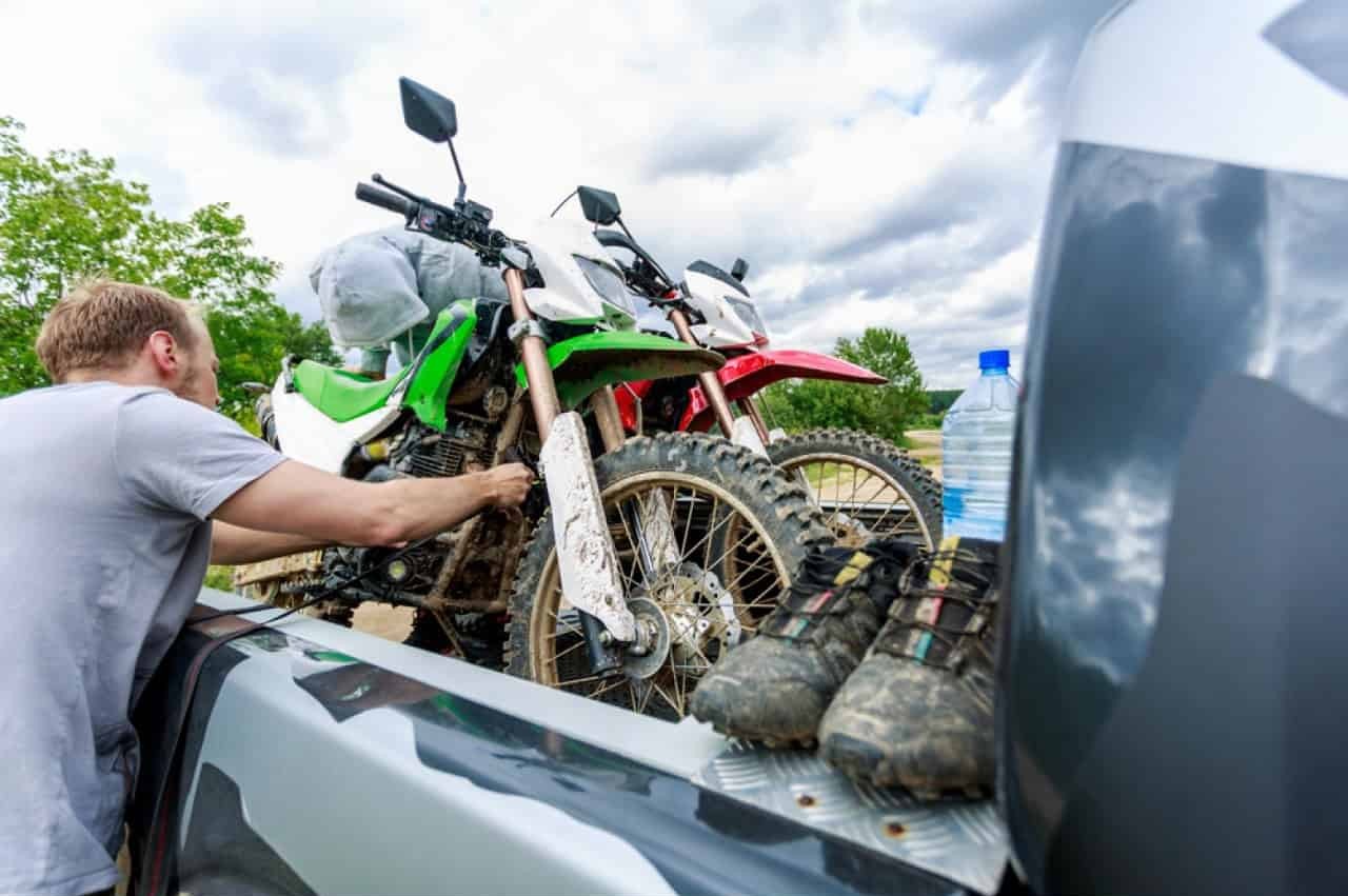 Man tightening a tie-down strap on a dirt bike to slightly compress the front suspension in the bed of a pickup truck