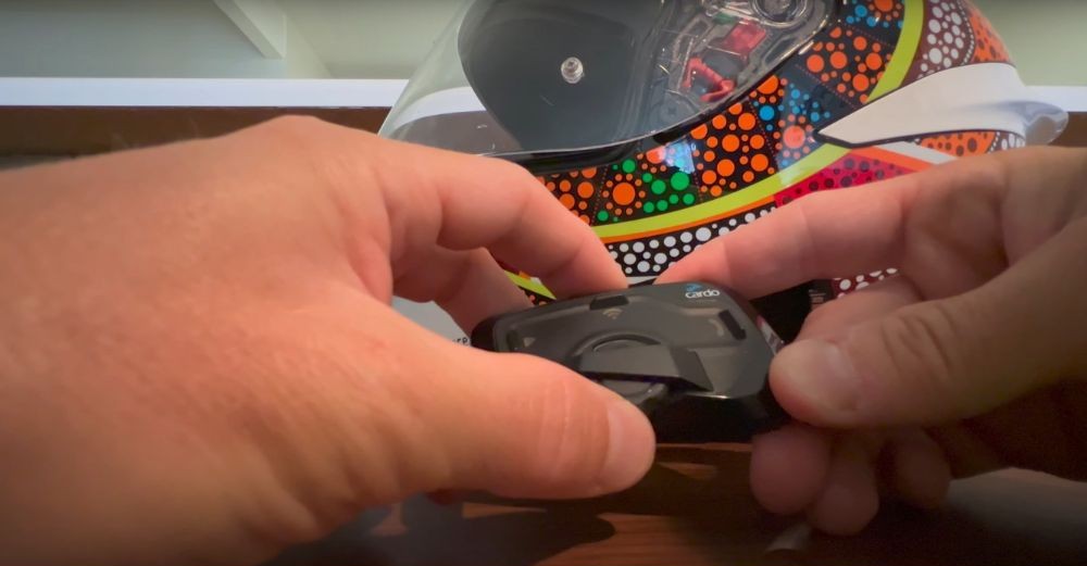 Hands adjusting the control wheel on a Cardo Freecom 4X unit next to a colorful motorcycle helmet.