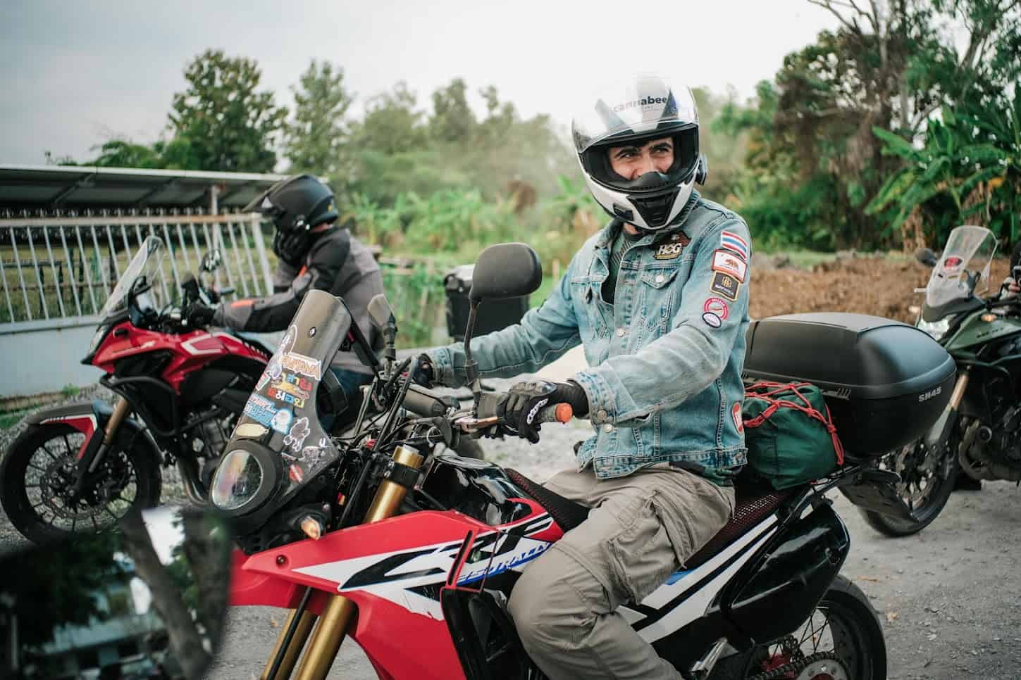 Motorcyclist leading a group ride, wearing a white helmet with Cardo unit, denim jacket, and gloves, preparing to roll out on a red ADV bike.
