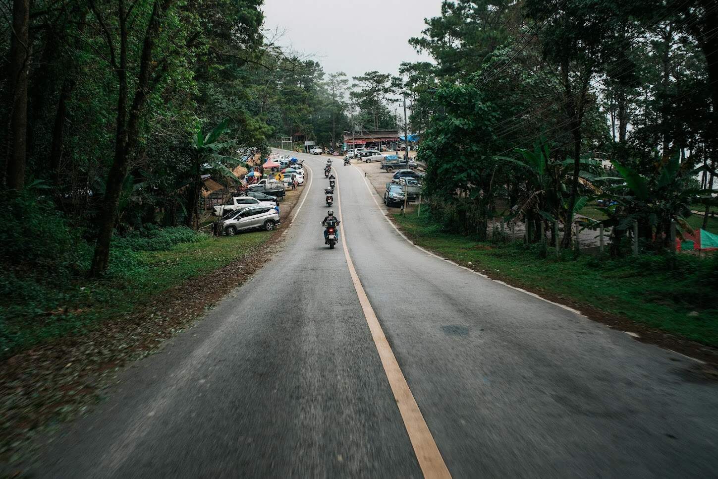 Motorcycle group riding in formation through a rural road surrounded by forest and roadside stalls during a tour.