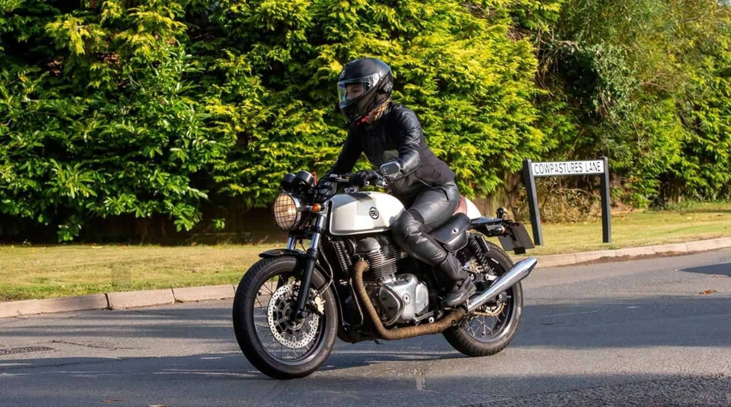 Woman riding a motorcycle in full gear including helmet, leather jacket, pants, and motorcycle boots, turning on a paved street near greenery.