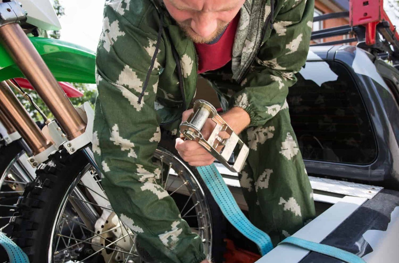 Man tightening a blue ratchet strap to secure a dirt bike in the back of a pickup truck