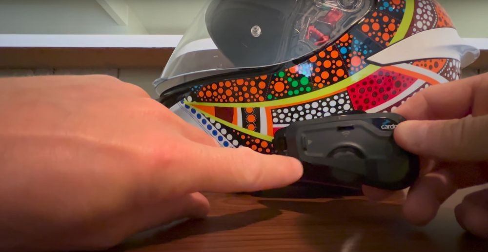 Close-up of hands demonstrating the Cardo Freecom 4X release button next to a helmet with a colorful design.