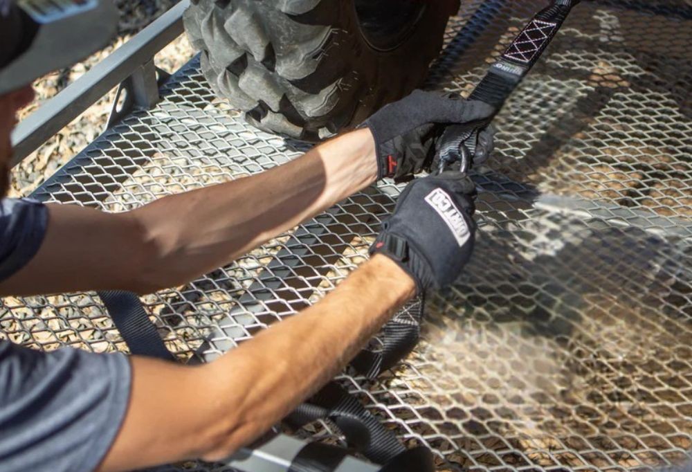 Person tightening a heavy-duty tie-down strap with gloves on a mesh trailer deck, securing an off-road vehicle