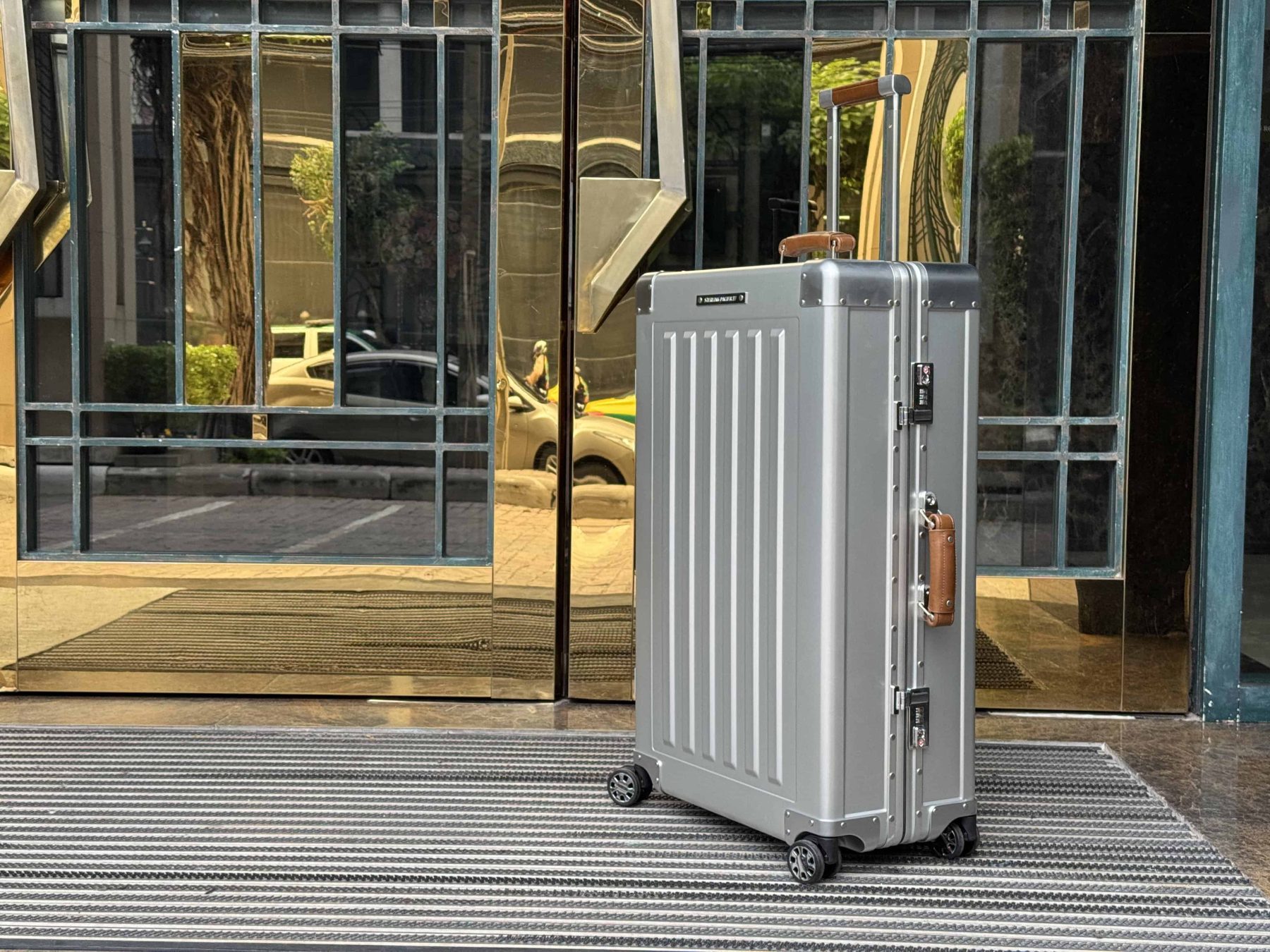 Sterling Pacific aluminum suitcase standing outside a reflective golden hotel entrance, showcasing its timeless design and premium finish.