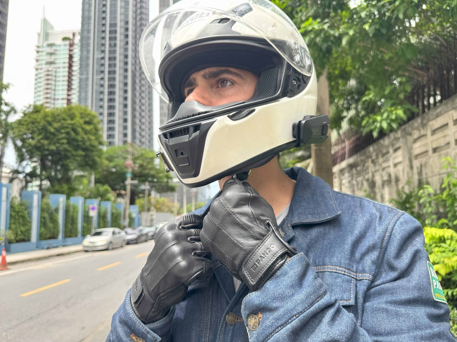Motorcyclist wearing a white helmet with Cardo unit and gloves, preparing for a ride on a city street with high-rise buildings in background.
