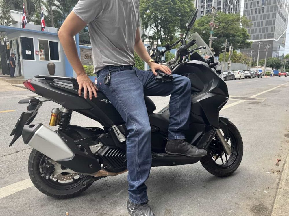 Man in casual riding gear sitting on a black scooter in an urban setting