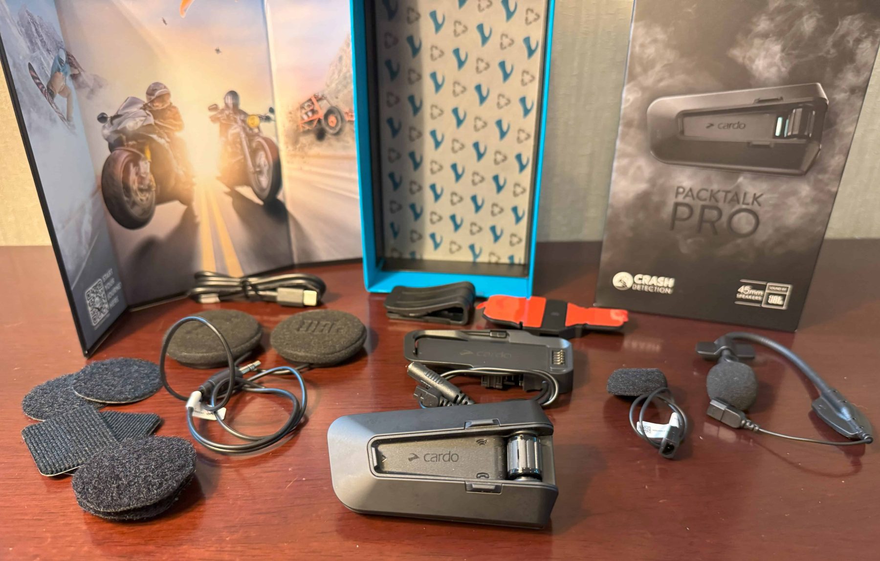 Cardo Packtalk Pro full kit laid out on a table, including the main unit, JBL speakers, microphones, and accessories with branded packaging.