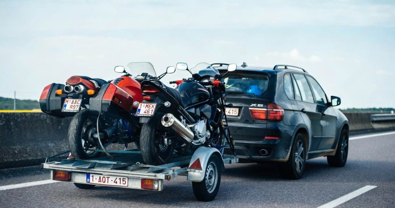 Two motorcycles secured with straps on a trailer behind a BMW SUV traveling at highway speed
