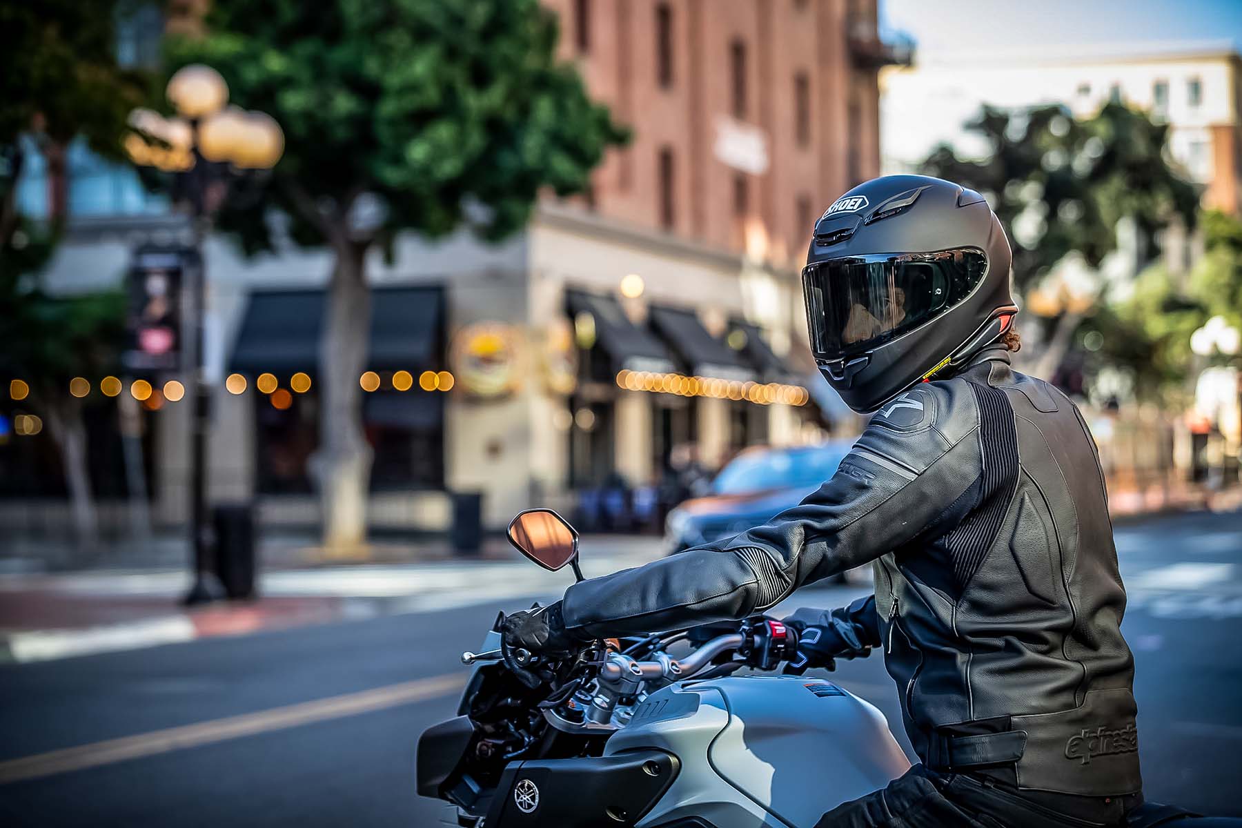 Motorcyclist wearing a matte black Shoei RF-1400 helmet and leather jacket, riding a sportbike through an urban city street.
