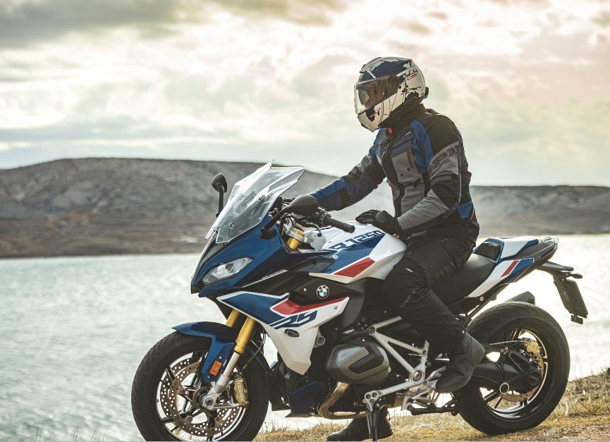 Rider on a BMW R1250RS wearing a touring helmet beside a lake at sunset