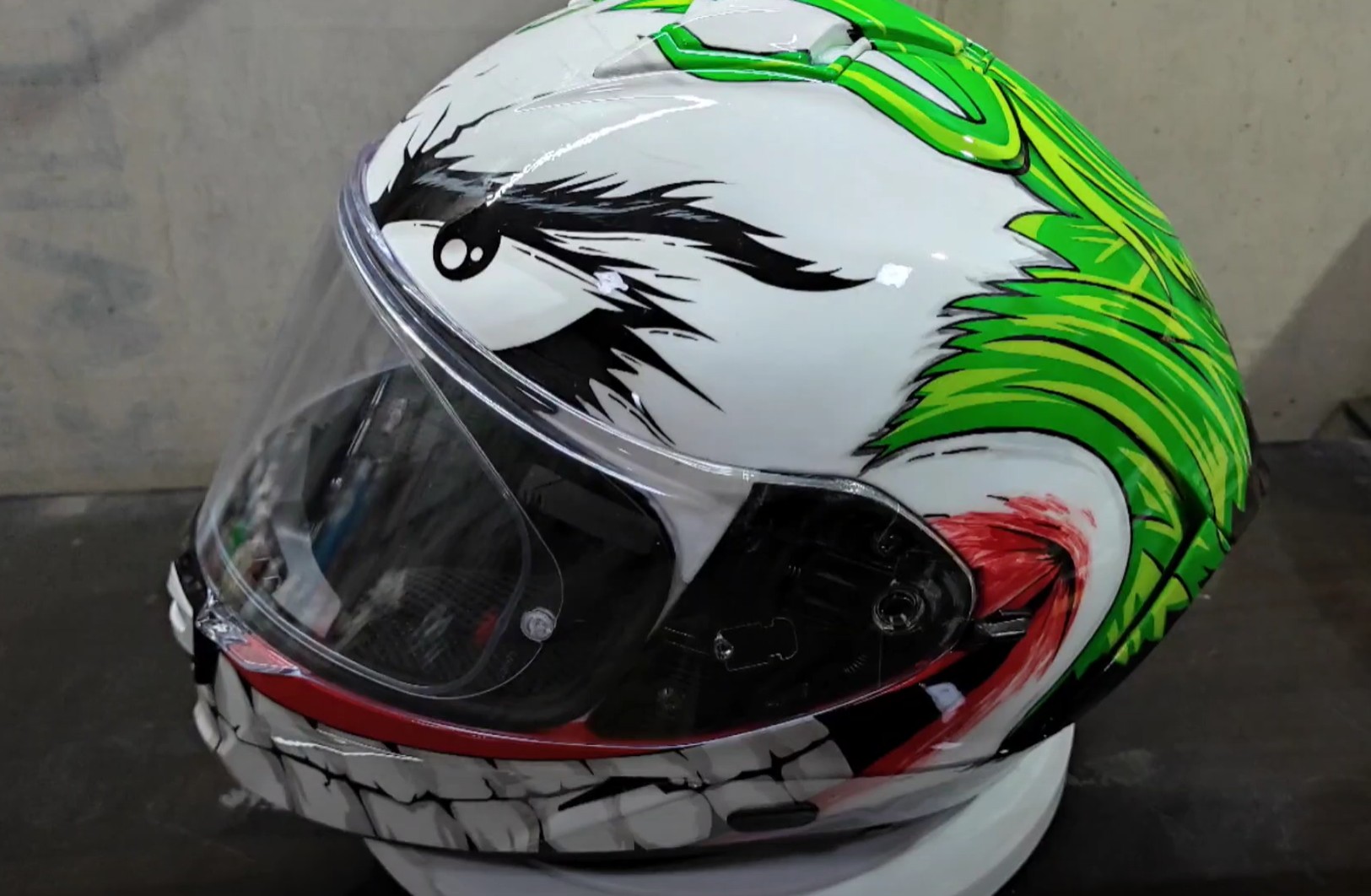 Side view of a full-face motorcycle helmet custom-painted with Joker-inspired graphics, including green hair, a sinister eye, and a wide grin.
