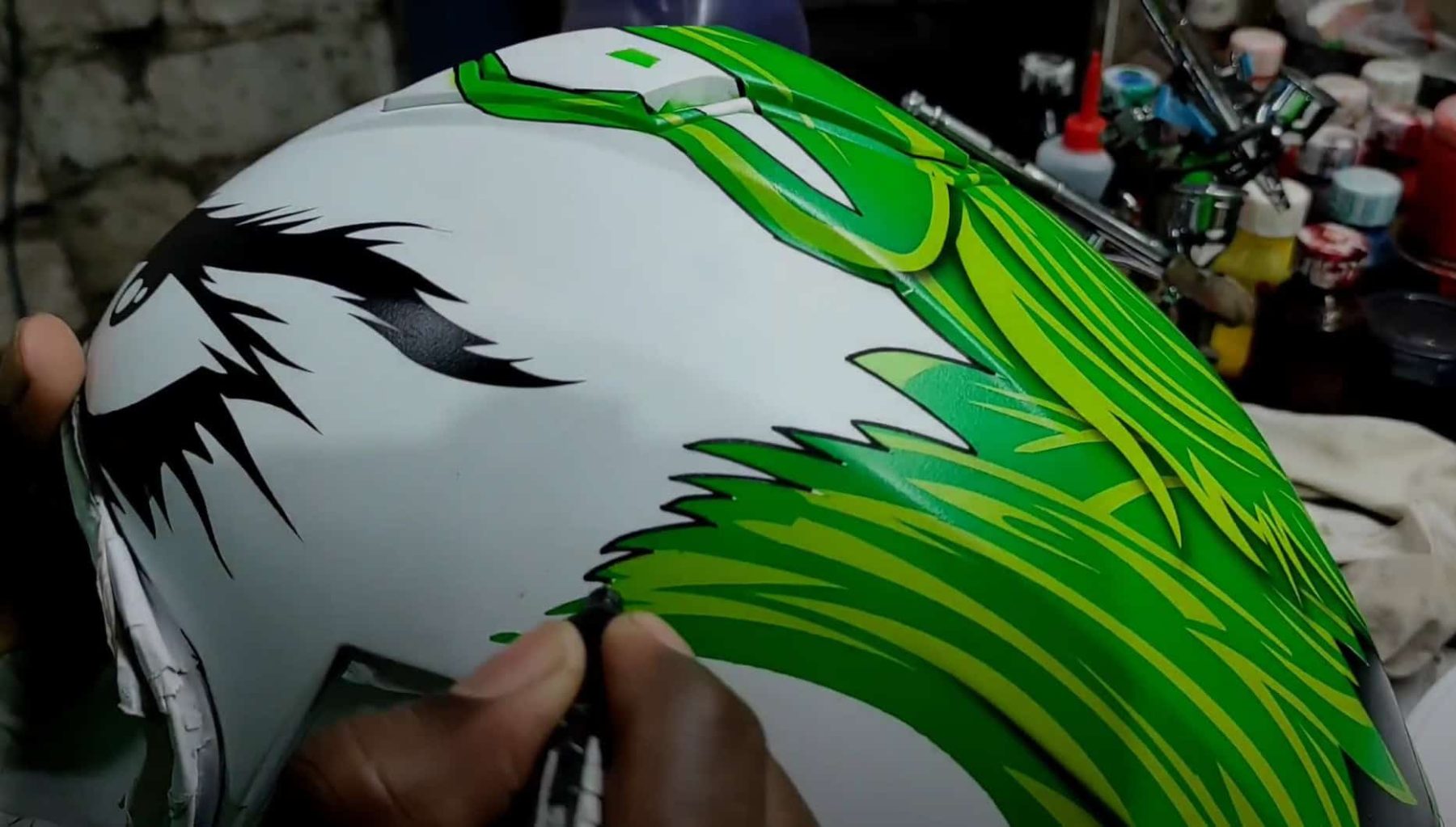 Close-up of an artist painting vivid green Joker hair onto a white motorcycle helmet using a fine brush.