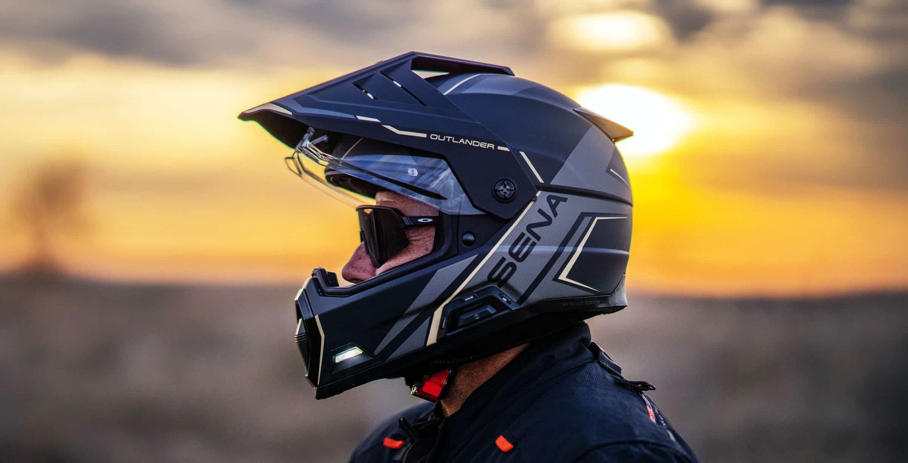 Rider wearing Sena Outlander Bluetooth helmet with peak visor and integrated lights at sunset