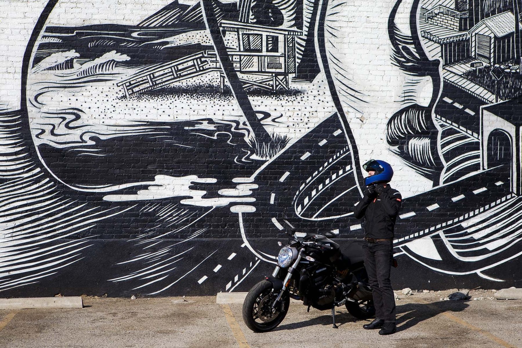 Motorcyclist gearing up in front of a mural with a blue helmet and a black street bike