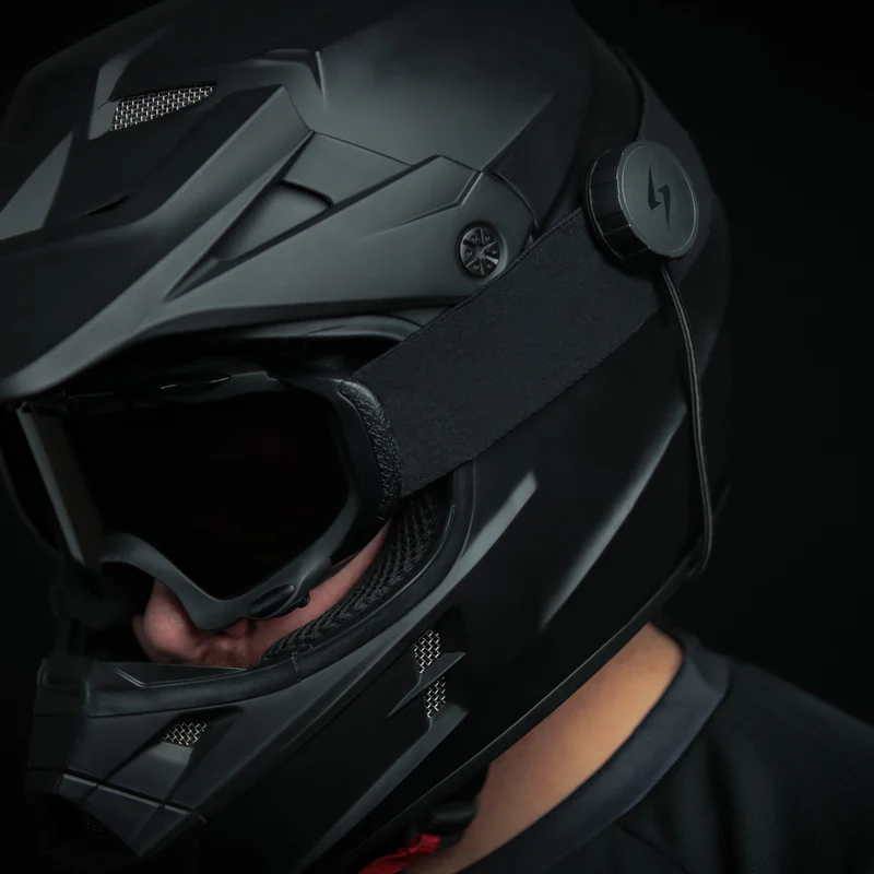 Close-up of a motorcyclist wearing a black full-face helmet with Syphon SoundPro Ultra Thin speaker mounted externally on the goggle strap.