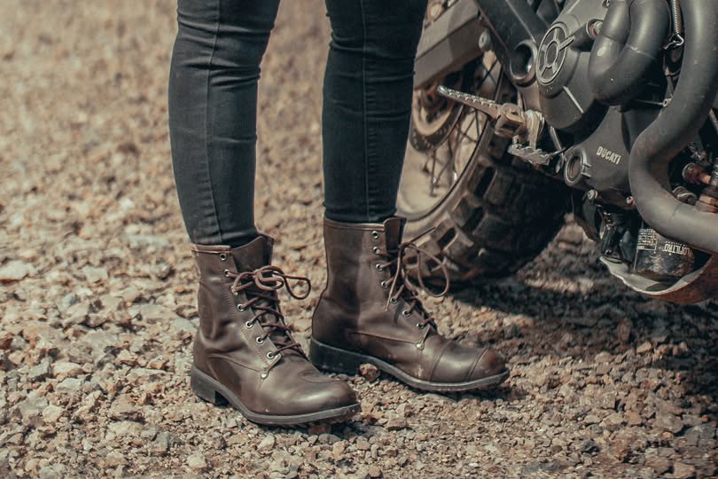 Close-up of a woman wearing brown lace-up motorcycle boots standing beside a Ducati bike on gravel, showcasing proper fit and riding gear.