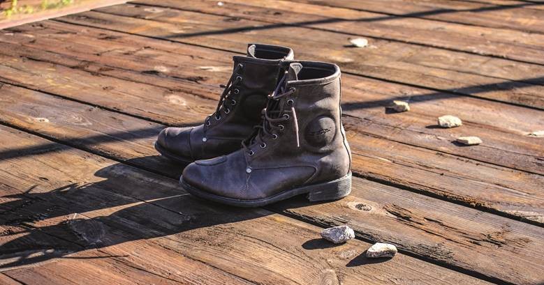 A pair of dark brown lace-up motorcycle boots resting on a sunlit wooden deck, showing rugged construction and worn-in character.