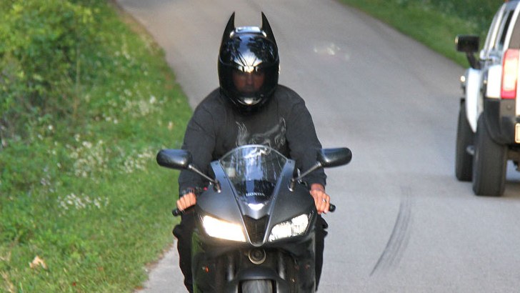 Motorcyclist wearing a black Batman-style helmet with pointed ears, riding a sportbike down a country road in daylight.