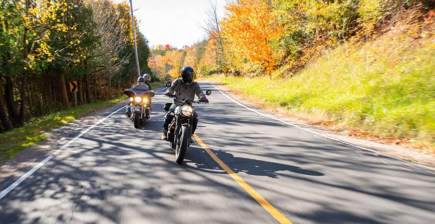 Two motorcyclists ride through fall foliage on a rural Canadian highway.