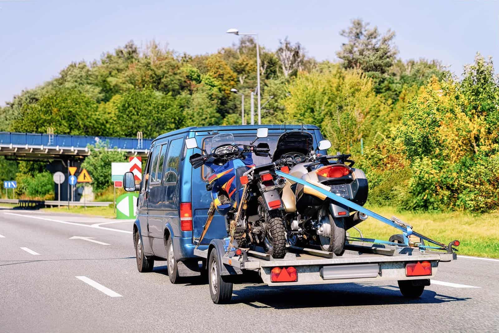 Two motorcycles strapped to a trailer behind a blue van traveling on a highway