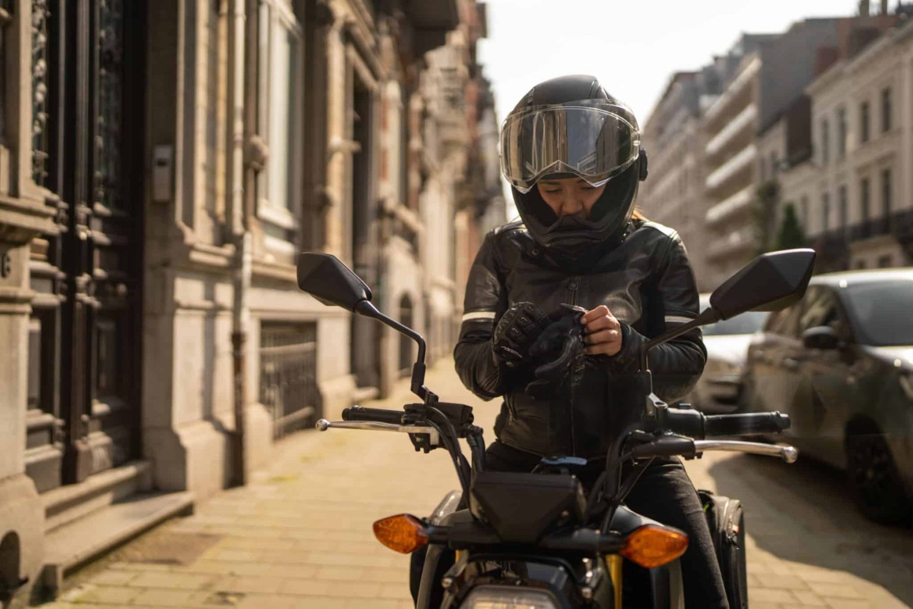 Woman in a black helmet and leather jacket sitting on a motorcycle, adjusting her gloves on a city street.