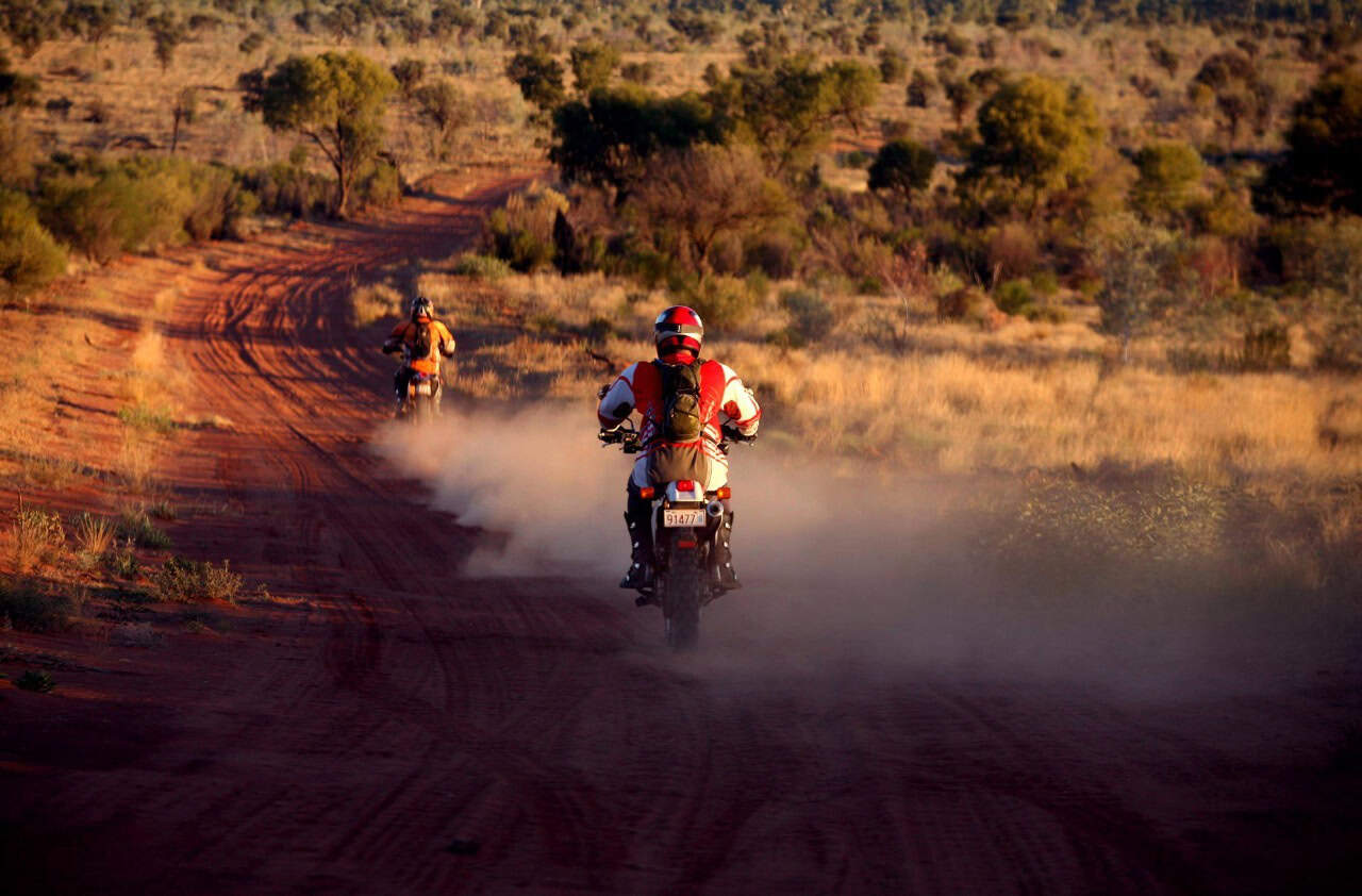 Two motorcyclists riding off-road through red Outback dirt, kicking up dust at sunset