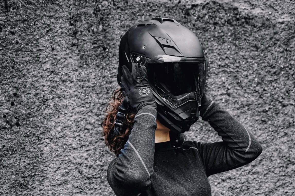 Woman adjusting a matte black motorcycle helmet against a textured wall