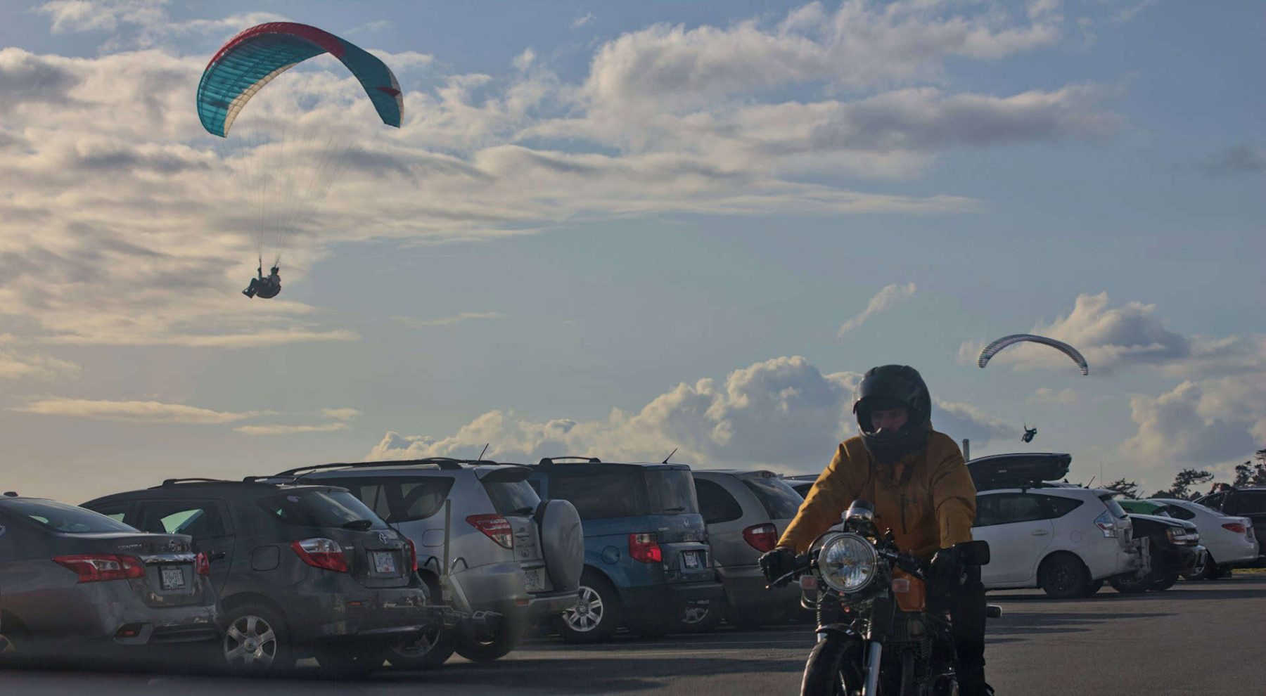 Motorcyclist wearing a helmet rides past parked cars as two paragliders soar in the background
