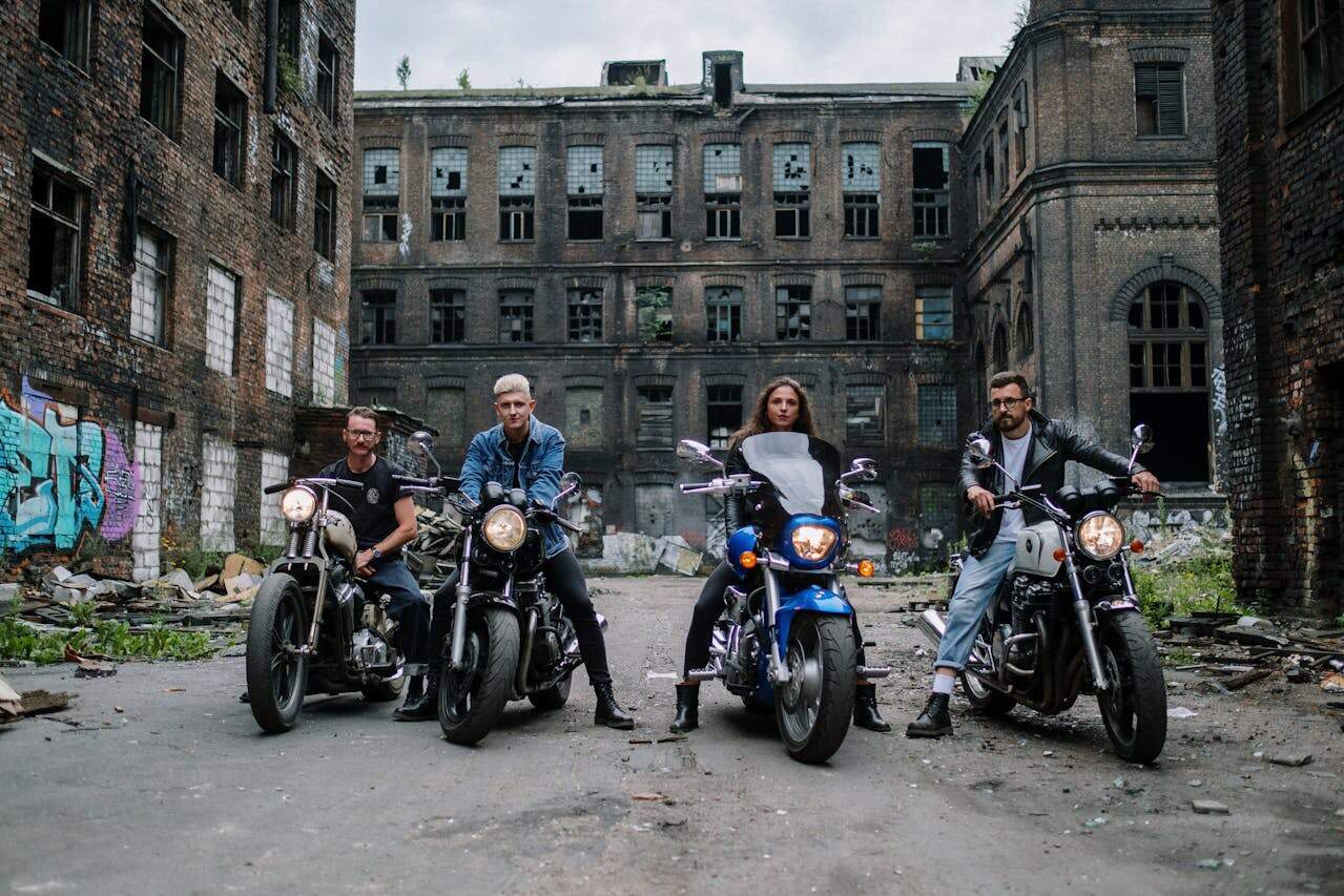 Group of four motorcyclists, including a woman, parked in front of an abandoned brick building, showcasing cruiser bikes and riding gear.