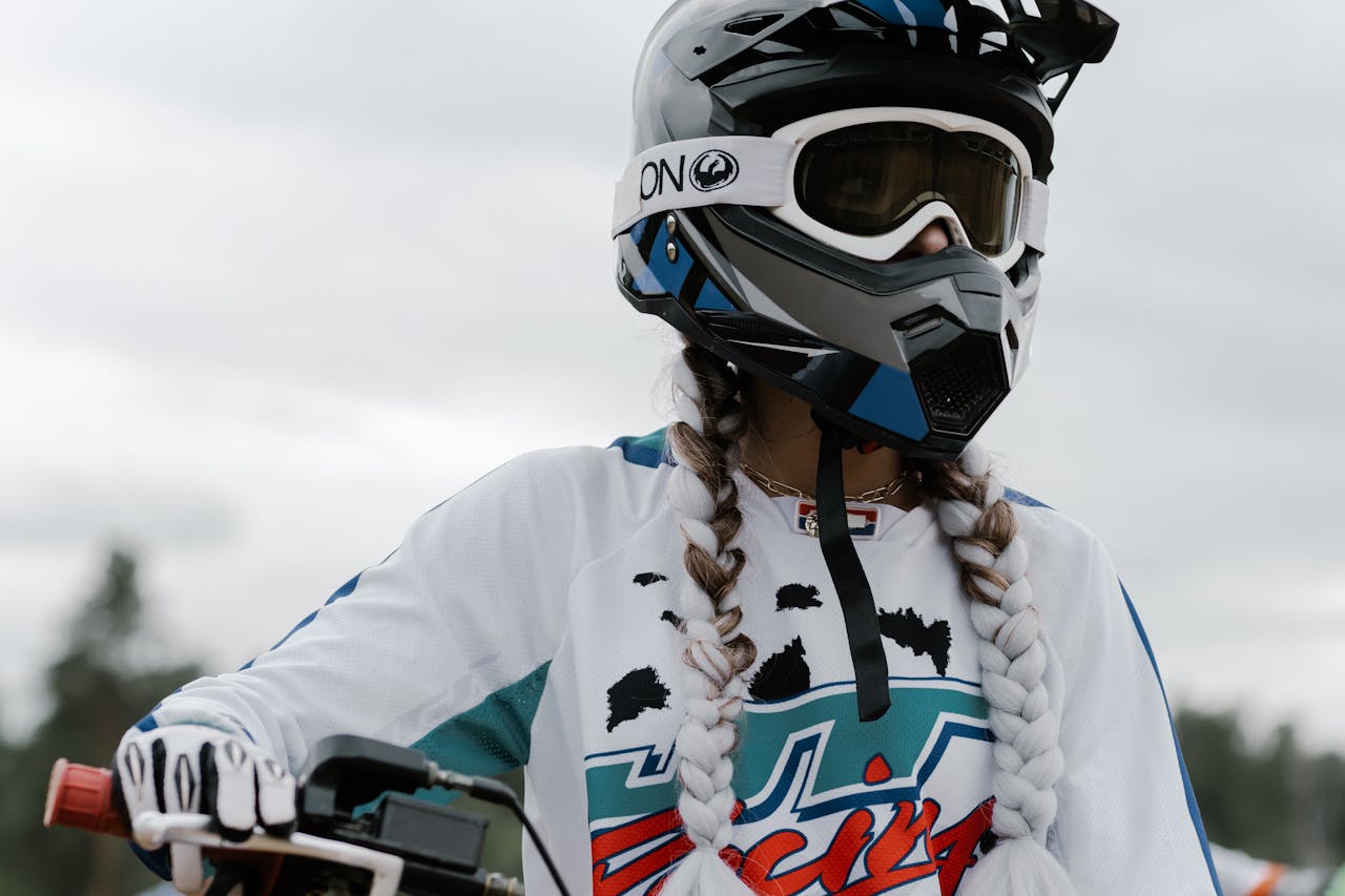 Woman wearing a motocross helmet with long twin braids, dressed in a white and blue racing jersey.