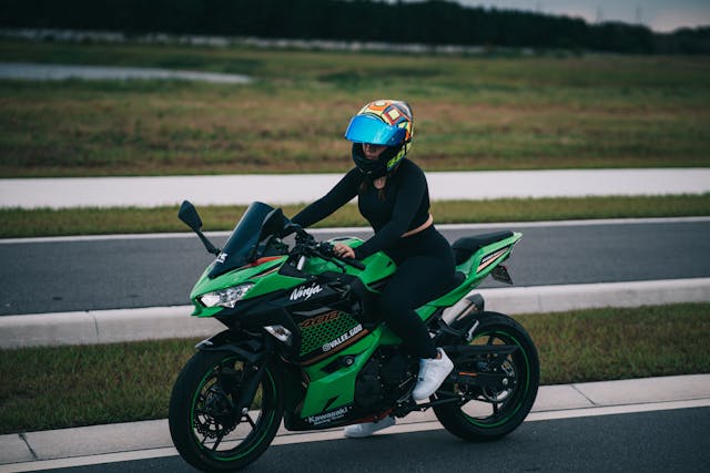 Woman riding a Kawasaki Ninja 400 in full gear, wearing a colorful full-face helmet.